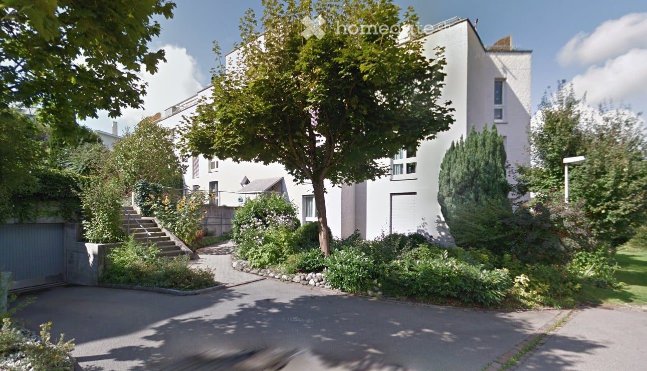 Modern white building with raised ground floor, garage, and landscaped garden with trees and shrubs.
