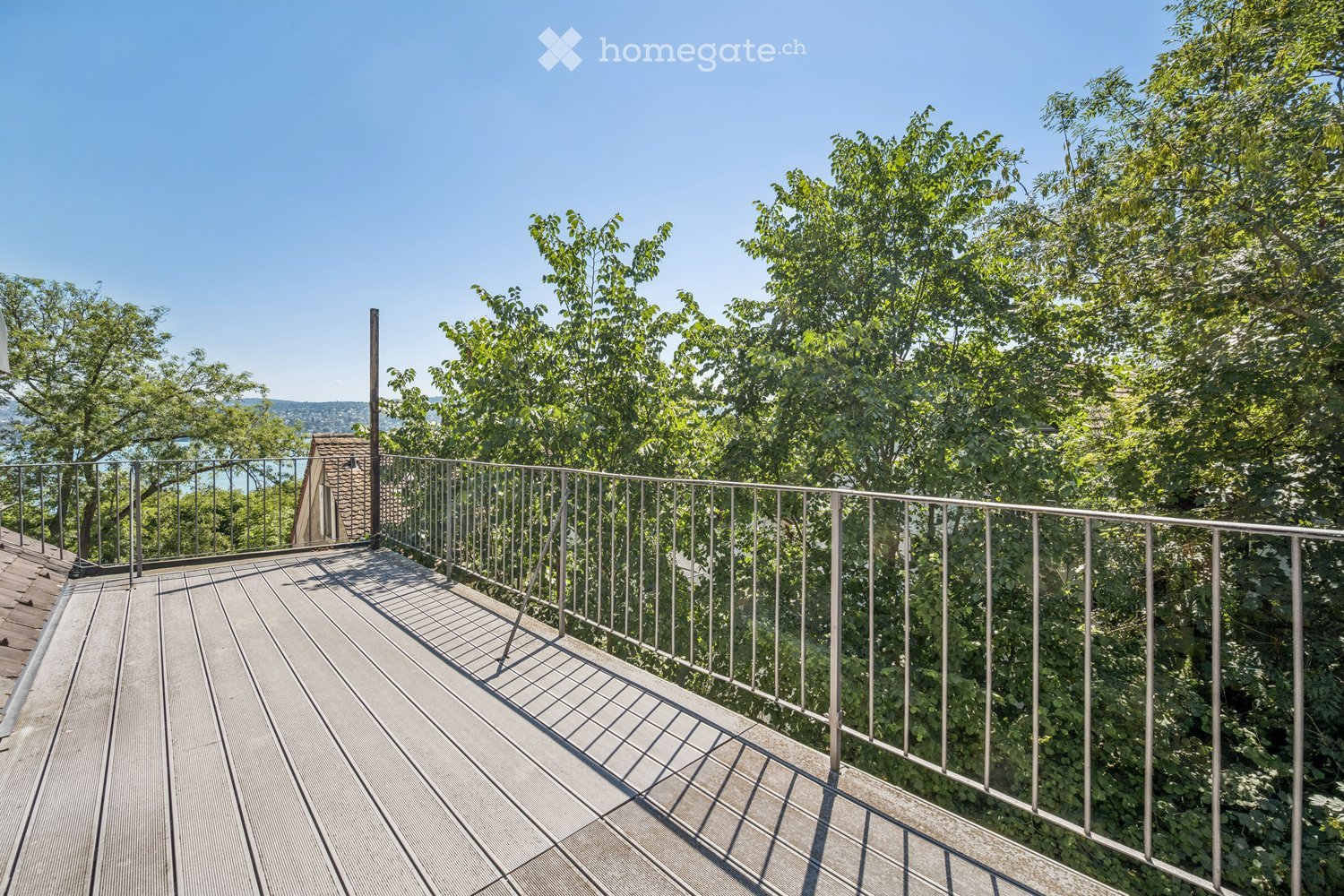 Wooden deck with metal railing, surrounded by lush green trees and a clear blue sky, offering a scenic view of the landscape.