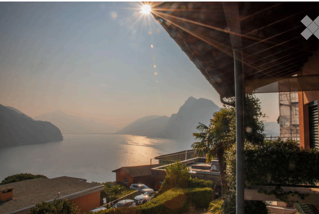 Balcony overlooking a serene lake with mountains in the background, surrounded by greenery and parked cars below.