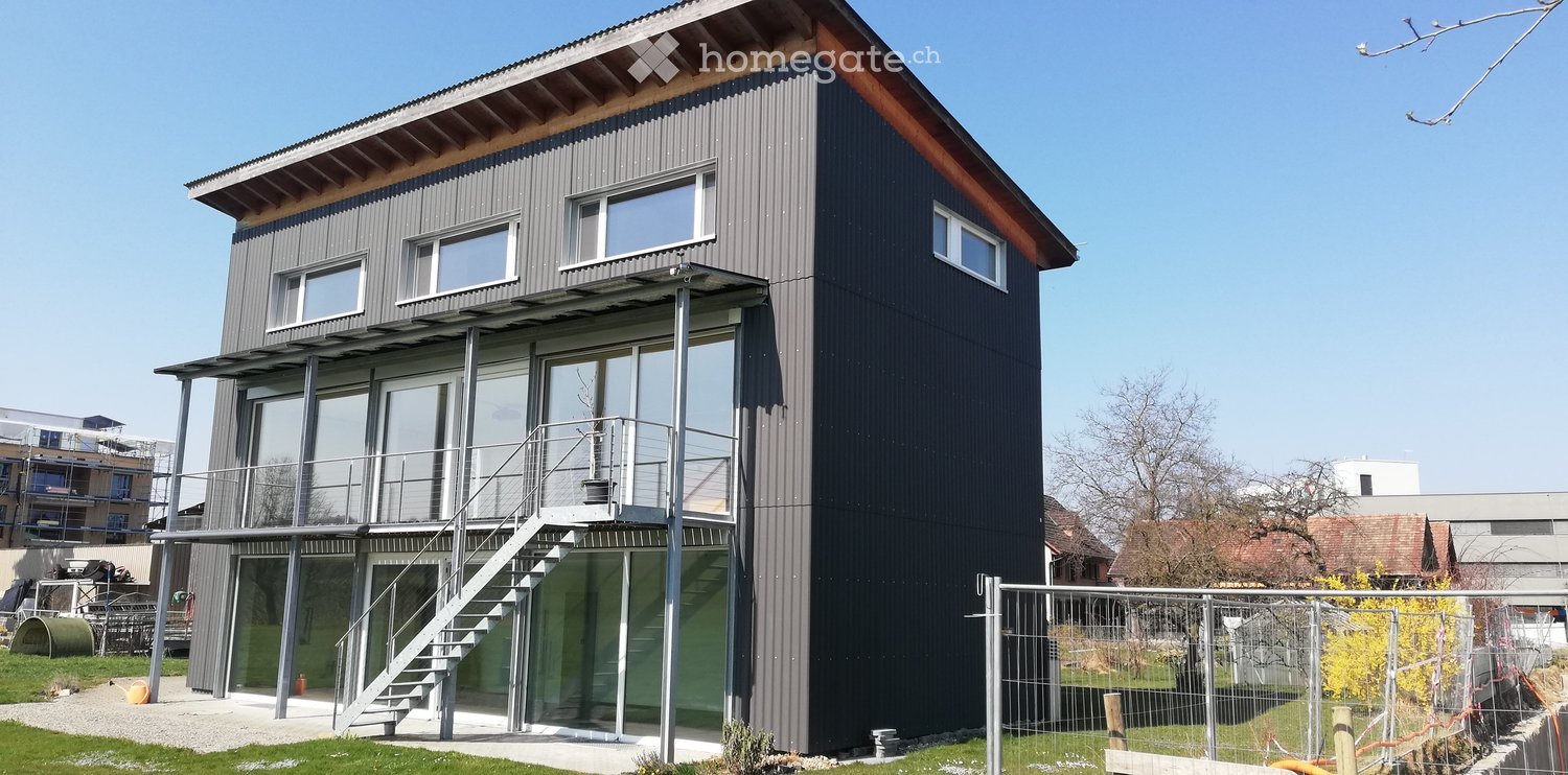 Modern two-story house with dark grey exterior, large windows, and a spacious balcony accessible from the upper floor.