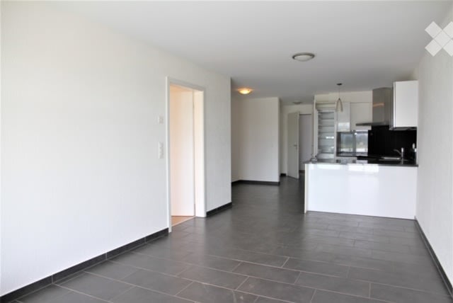 Empty, unfurnished room with white walls, dark tiled floor, and a doorway leading to another room. The adjacent room appears to be a kitchen with white cabinets and stainless steel appliances.