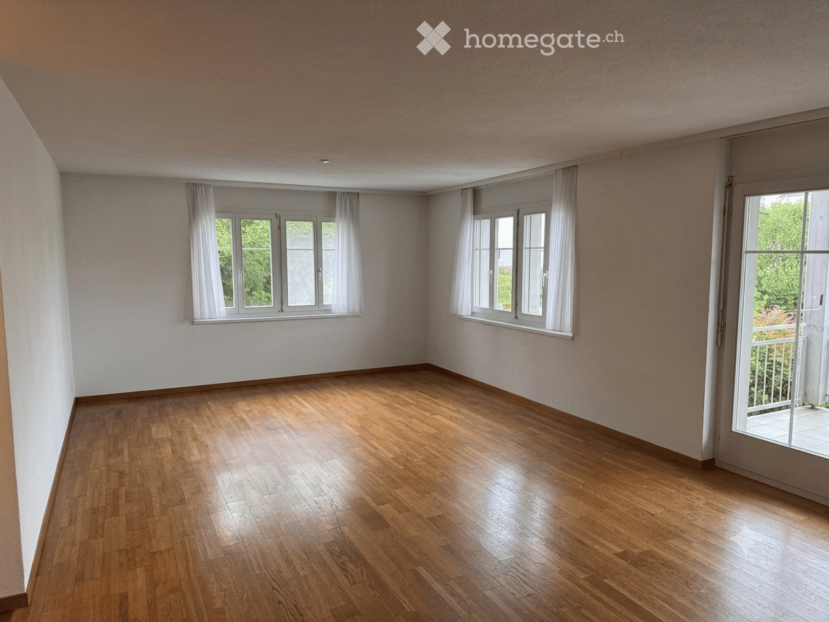 Empty room with wooden flooring, white walls, large windows with white curtains, and a sliding glass door leading to a balcony.