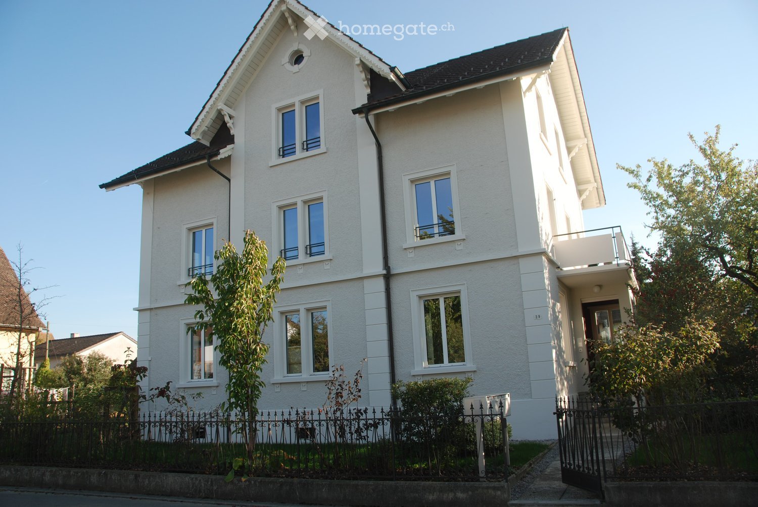 Two-story white house with multiple windows, a balcony, and a fenced garden area.