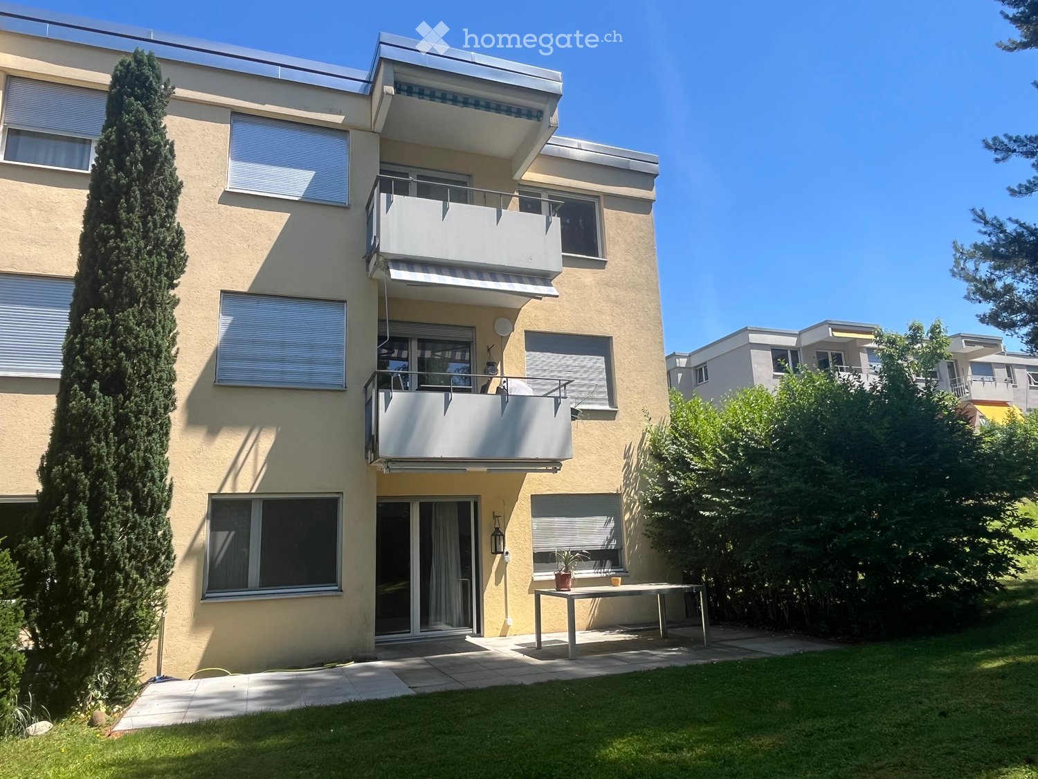 Modern two-story building with balconies, large windows, and a well-maintained garden area with a small table and potted plant.