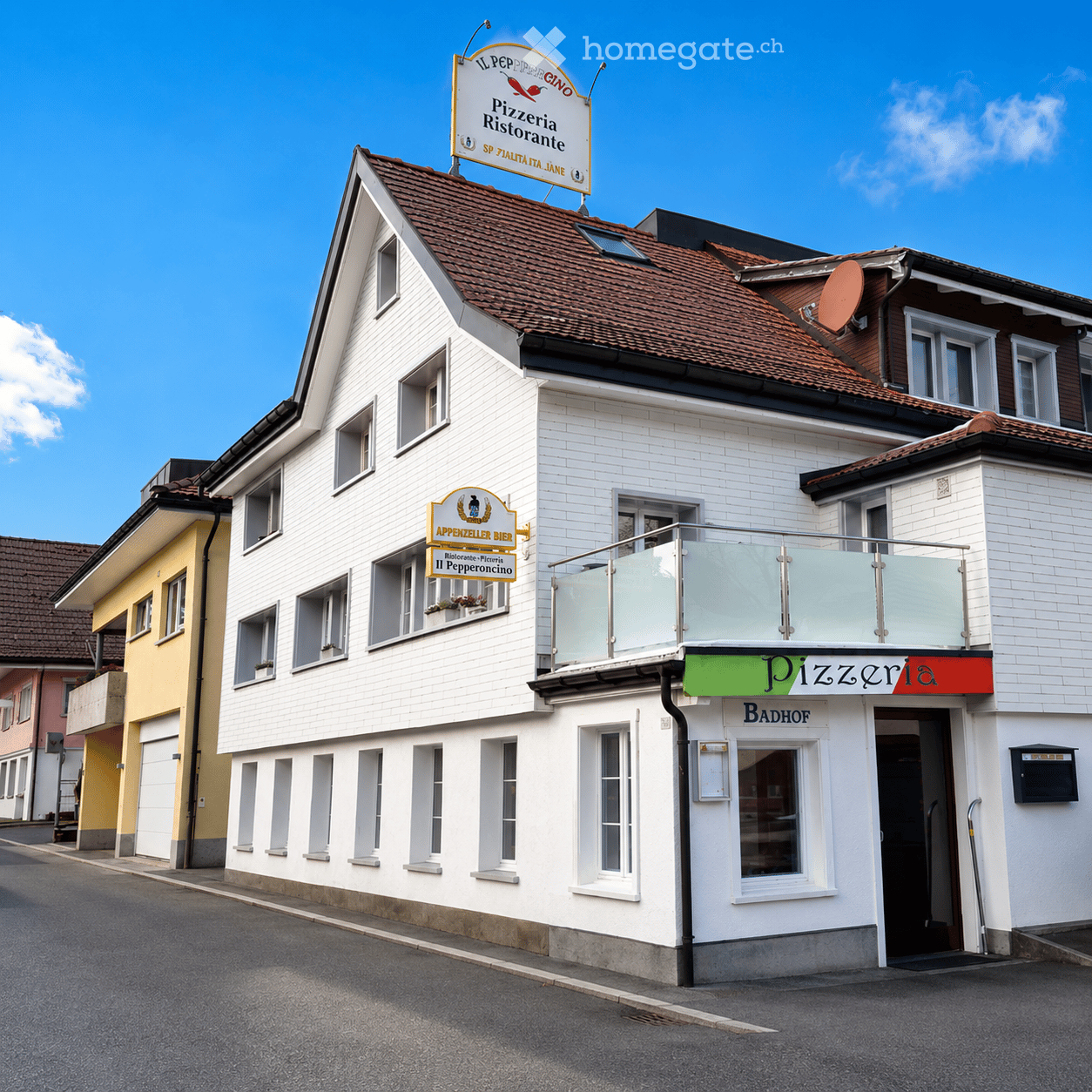 Two-story white building with a brown tiled roof, multiple windows, and signs indicating "Pizzeria Ristorante" and "Badhof". Features a balcony with glass railing and a satellite dish on the roof.