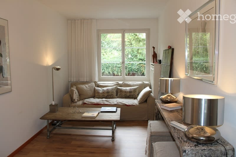 Living room with a beige sofa, coffee table, floor lamp, side table, and mirror. The room features large windows with curtains and a wooden floor.