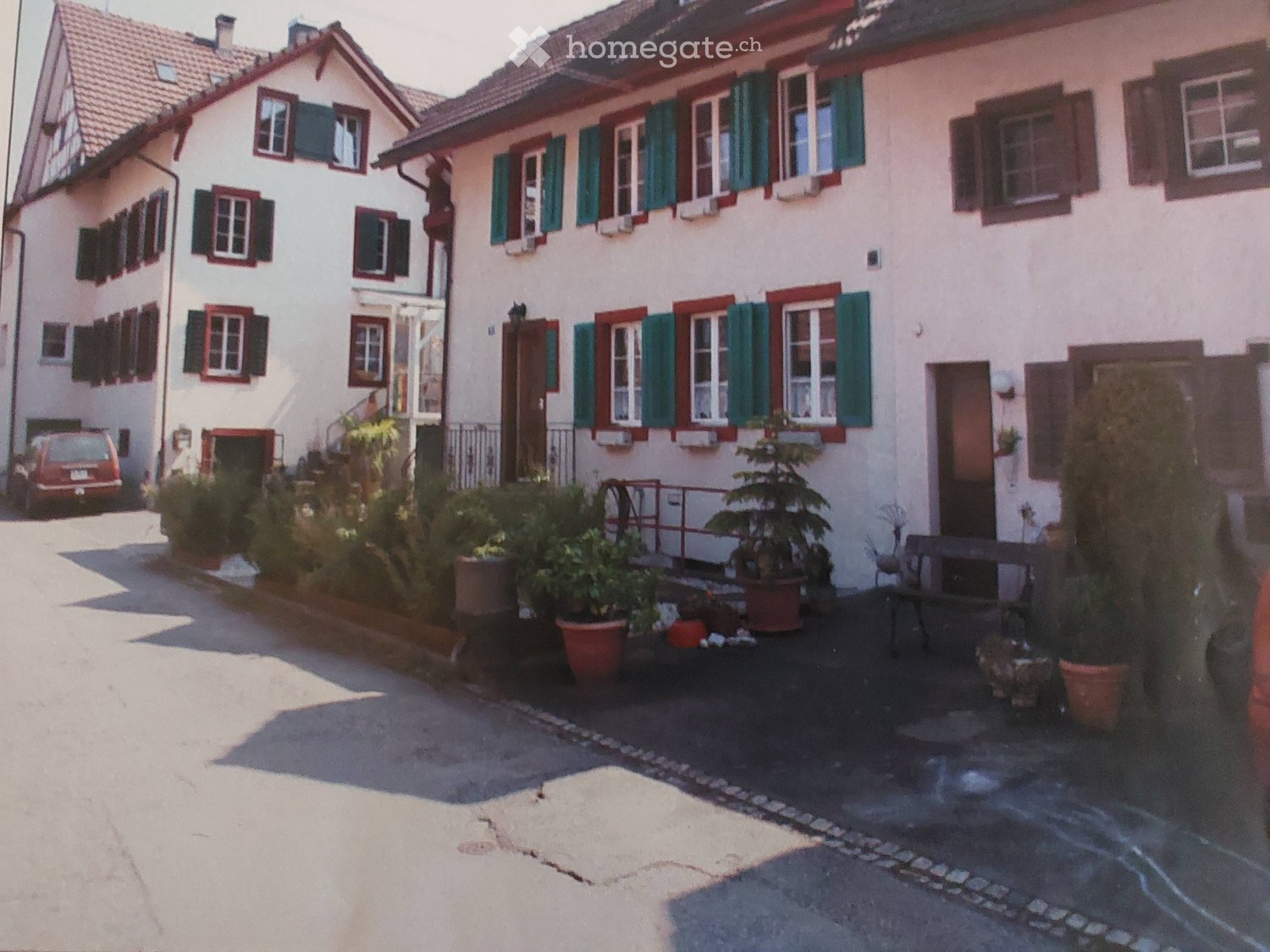 Two-story residential building with white walls, red shutters, and multiple windows. Potted plants and a bench are placed near the entrance.