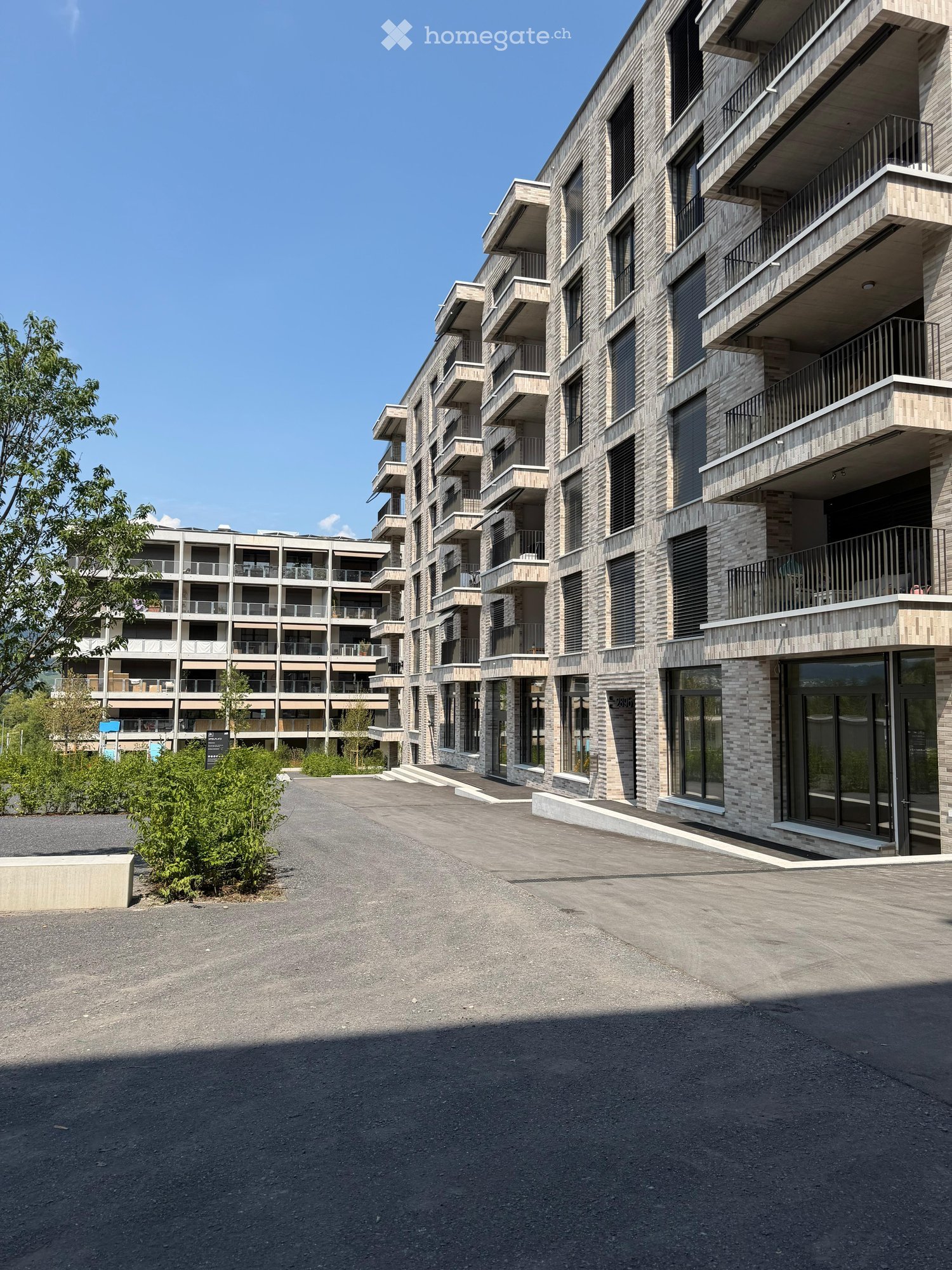 Modern multi-story residential building with balconies and large windows, surrounded by greenery and a paved area.