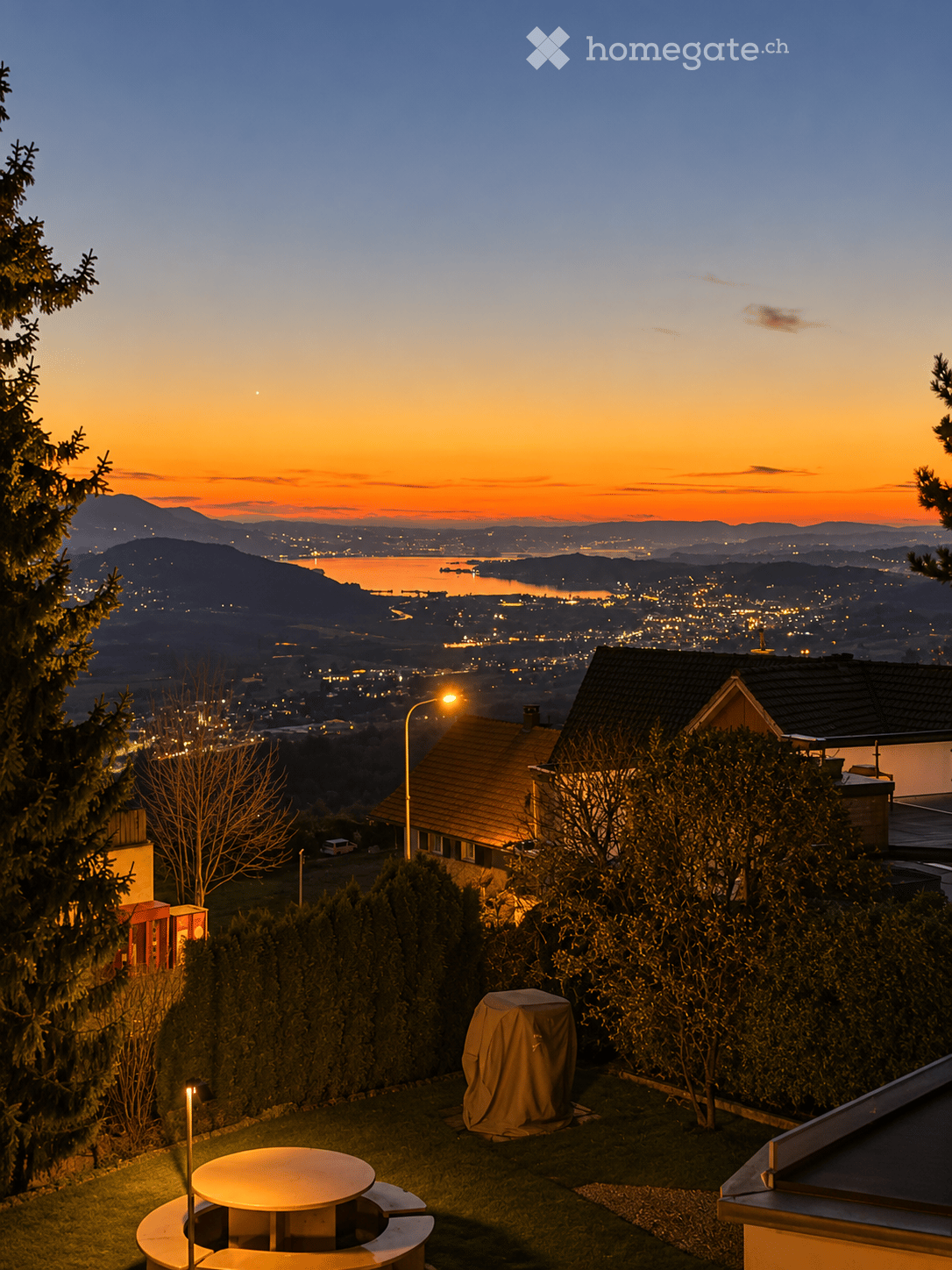 Garden with a round table and benches, overlooking a cityscape at sunset with mountains and a lake in the background.