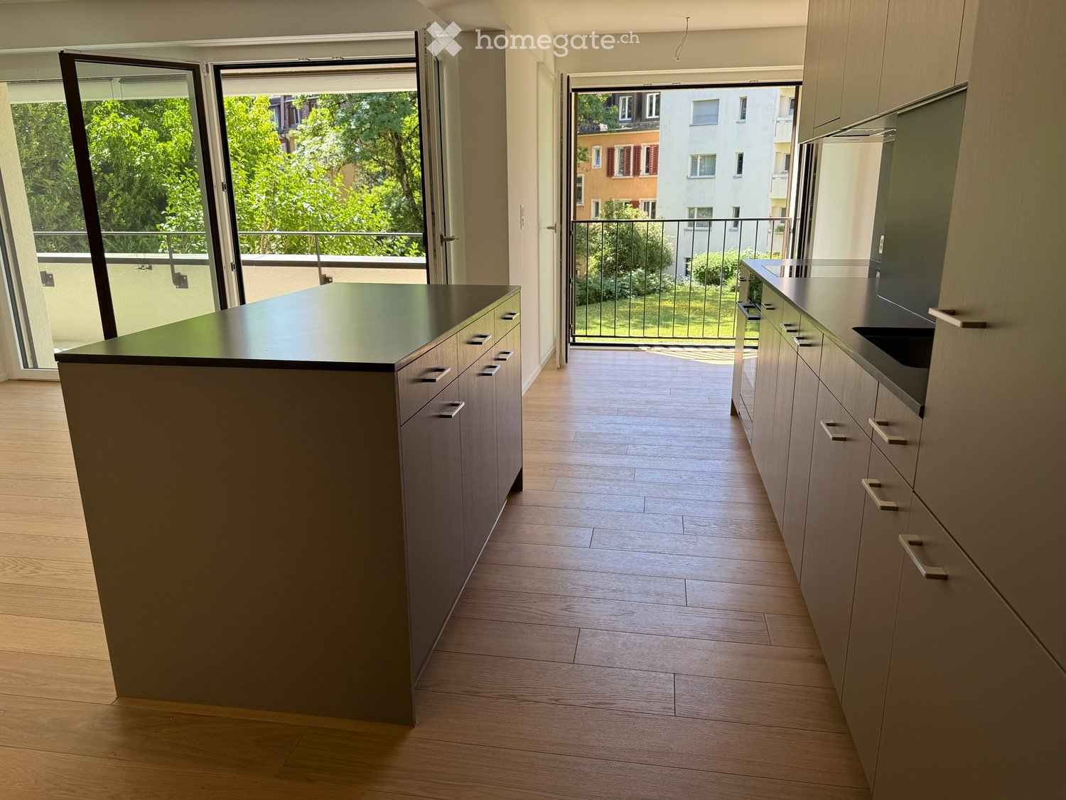 Modern kitchen with light wooden flooring, grey cabinets, and a central island with a black countertop. Large windows and sliding doors lead to a balcony with greenery visible outside.