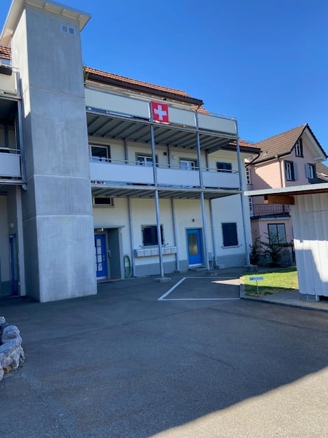 A modern multi-story building with a Swiss flag, featuring balconies, blue doors, and a concrete facade.