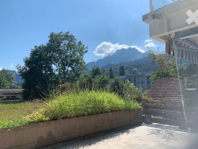 Balcony with a view of mountains and greenery, featuring a concrete planter with grass and a bench.