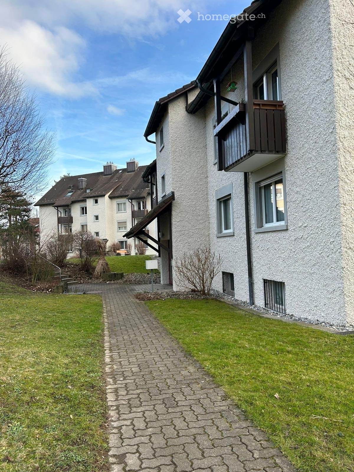 Two-story residential building with white facade, multiple windows, and a balcony on the upper floor. A paved pathway leads to the entrance.