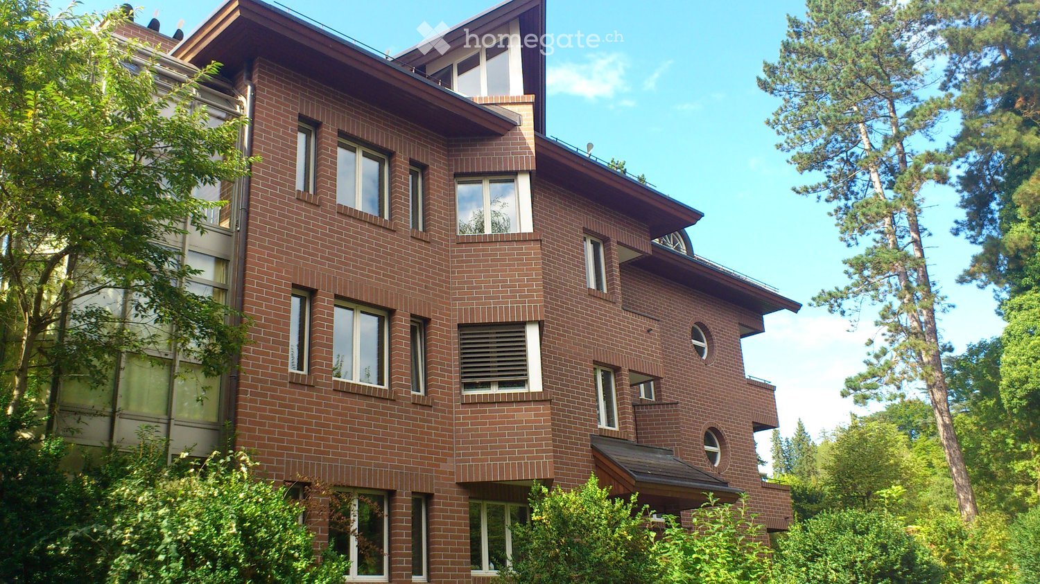 A multi-story brick building with large windows and a sloped roof, surrounded by trees and greenery.