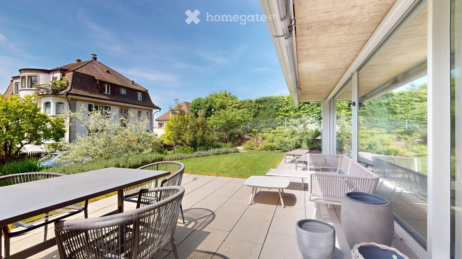 Balcony with a modern design, featuring a large table, several chairs, and a bench, overlooking a well-maintained garden and a residential building.