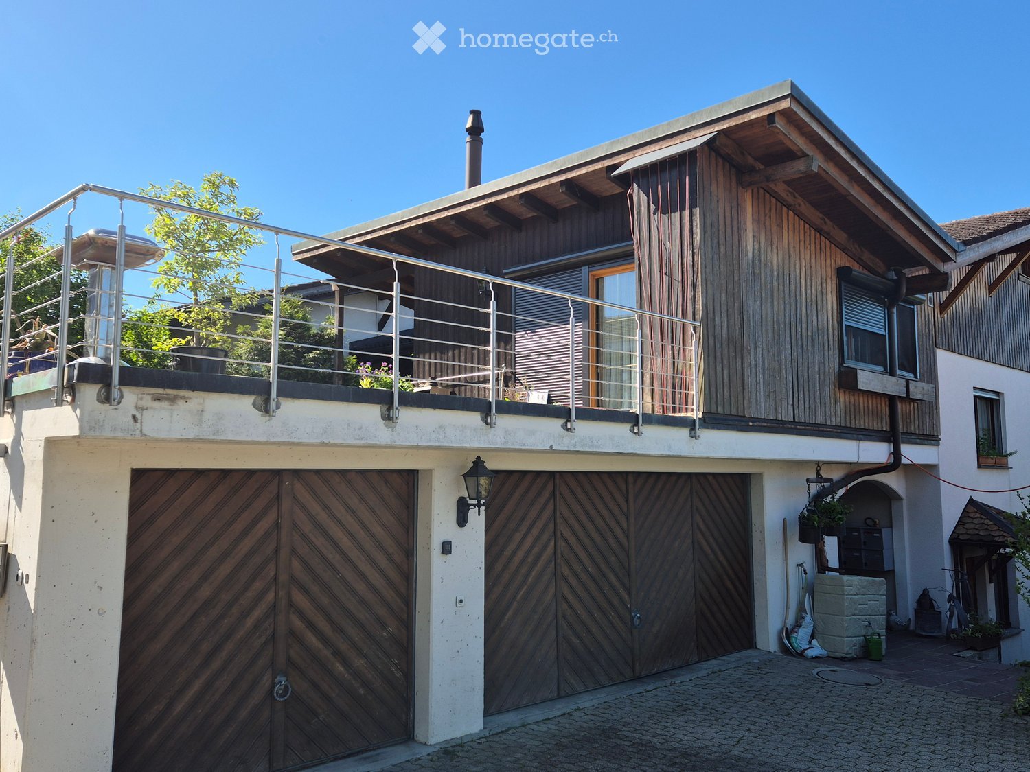 A residential house with wooden exterior walls, a metal railing balcony, a wooden garage, two black lanterns and two metal poles.