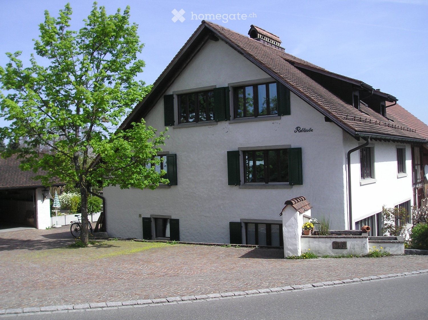 Two-story house with white walls, green shutters, and a brown tiled roof. Features include a garage, a driveway, and a small garden area with plants and a bicycle.