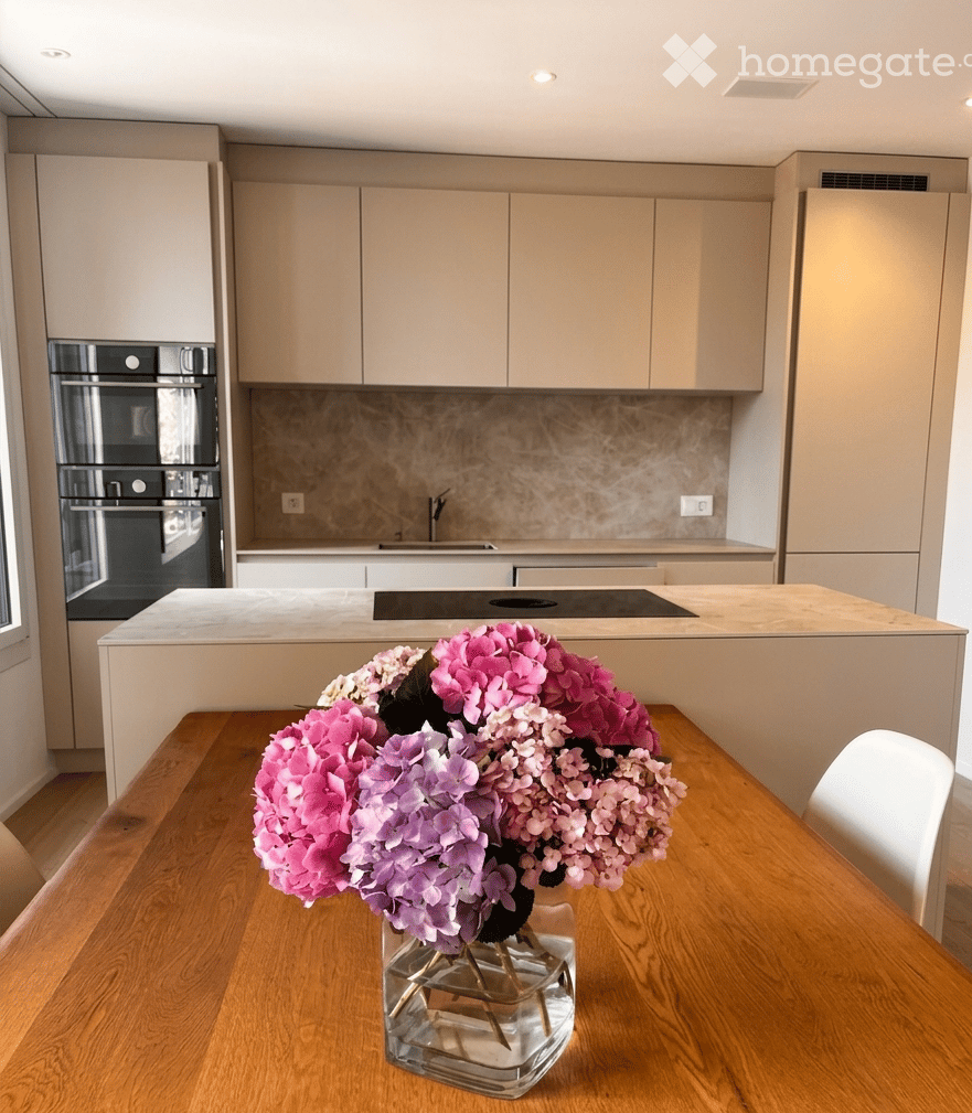 Modern kitchen with light-colored cabinets, a central island, and a vase of pink and purple flowers on the dining table.