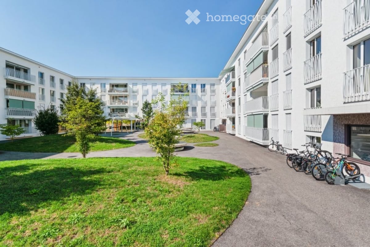 Modern multi-story residential building with white facades, balconies, and a landscaped courtyard with green spaces and pathways.
