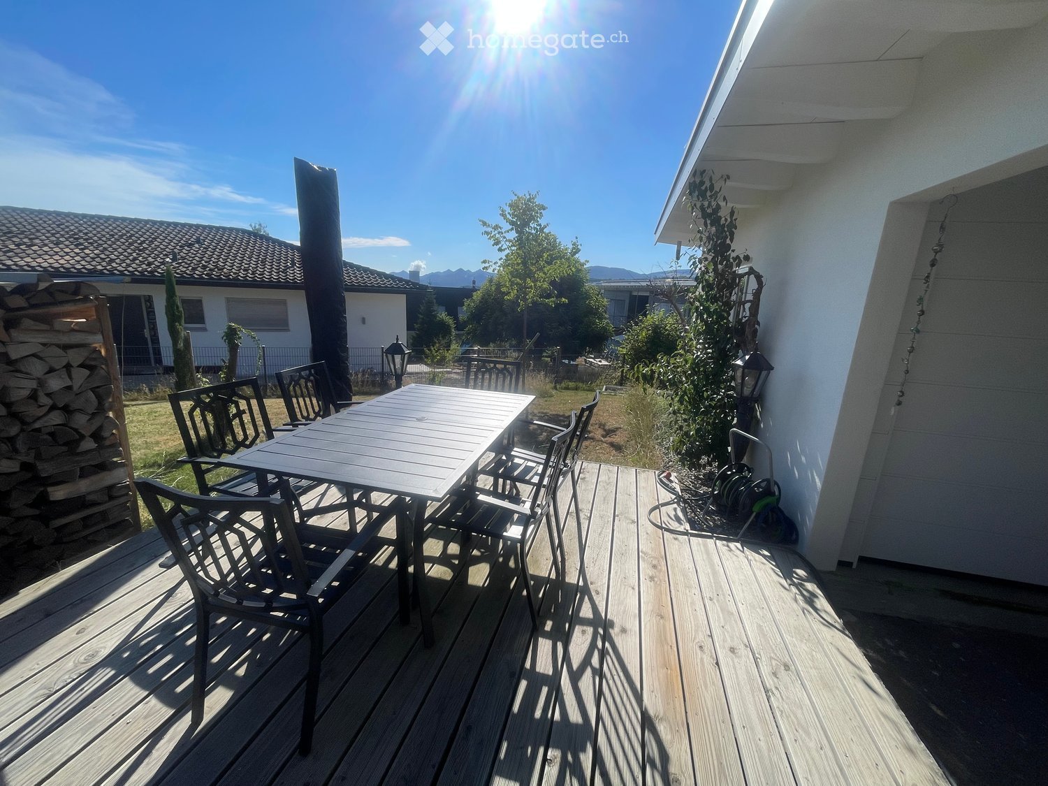 Wooden deck with a table and chairs, surrounded by a garden and neighboring houses.
