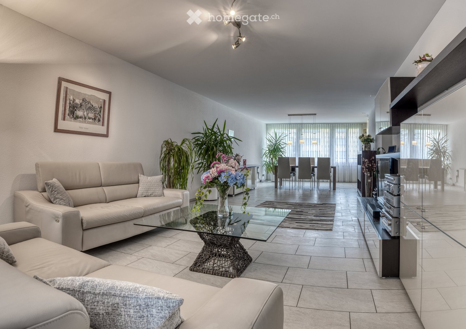 Modern living room with a beige sofa, glass coffee table, dining area with chairs and table, and decorative plants.