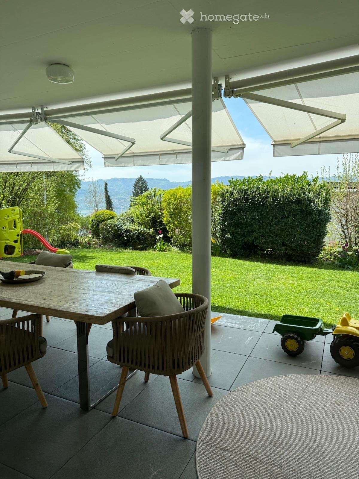 Outdoor patio with a wooden table, wicker chairs, and a view of a green lawn and distant mountains.
