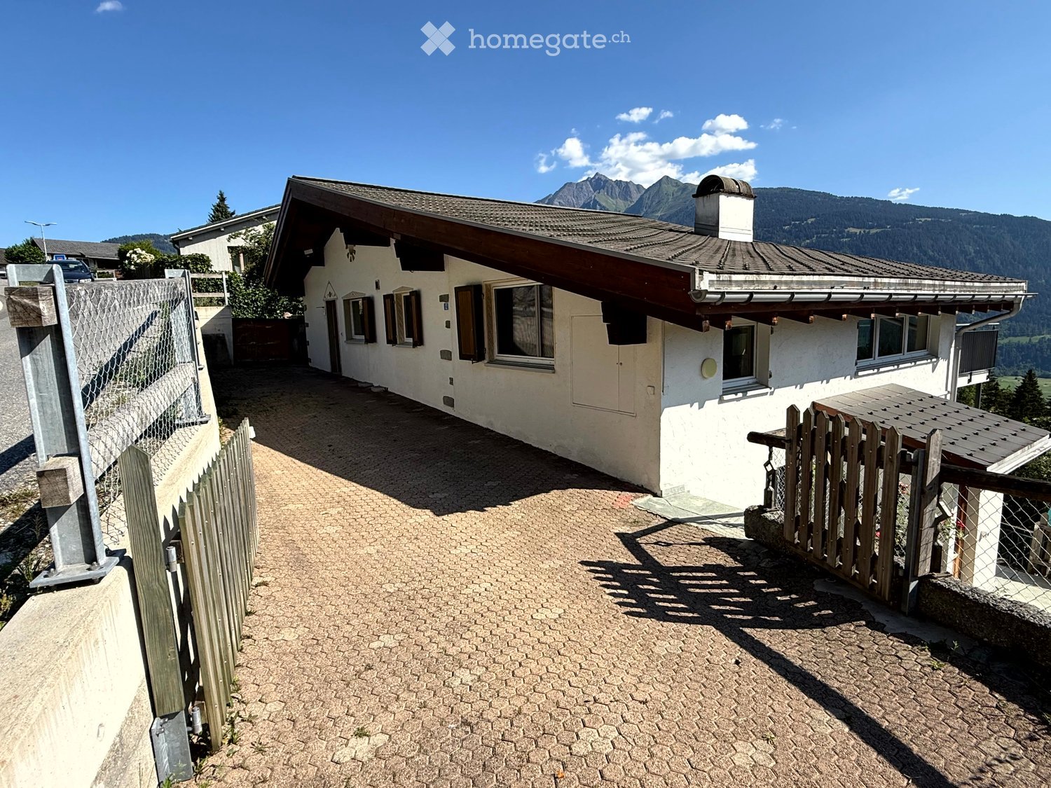 A two-story house with a white exterior and dark brown roof, featuring multiple windows and a chimney. The house is surrounded by a paved driveway and wooden fences, with a scenic mountain view in the background.