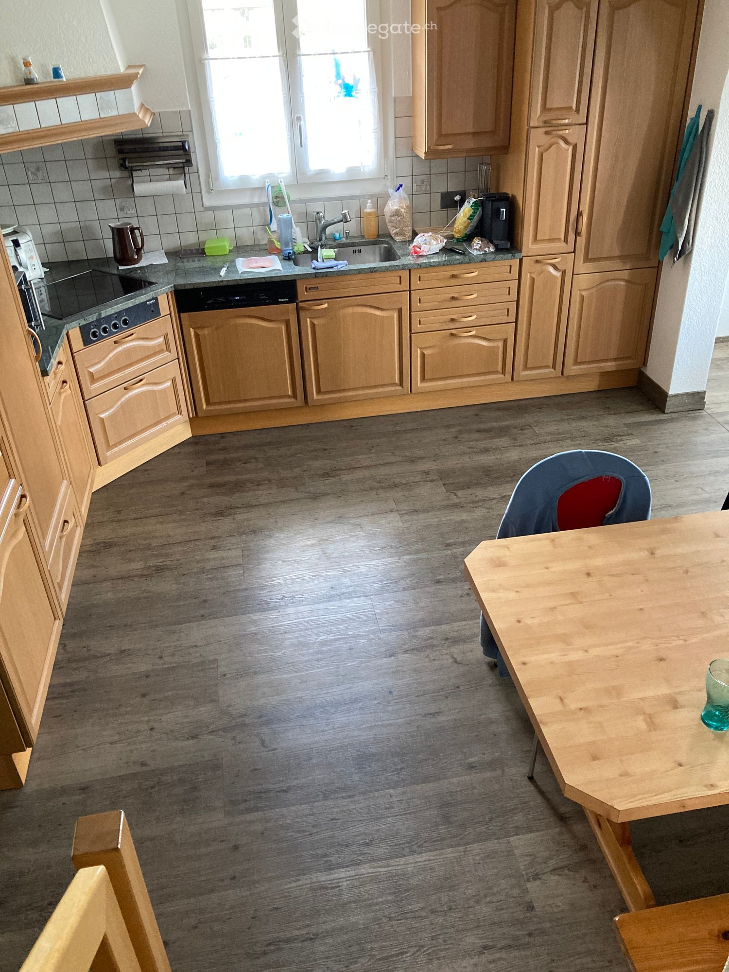 The kitchen features wooden cabinets, a granite countertop, a sink with a faucet, an oven, a dishwasher, a wooden dining table with a chair, and a glass on the table.