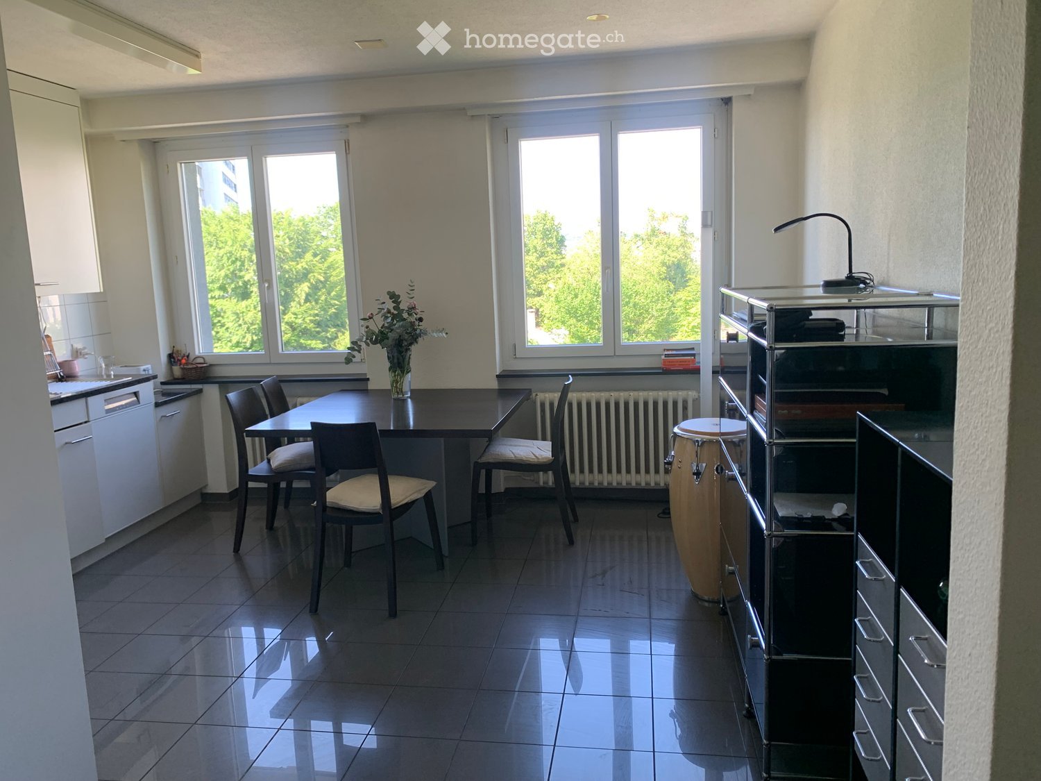 Bright room with large windows, tiled floor, dining table with chairs, vase with flowers, drum, and a modern cabinet.
