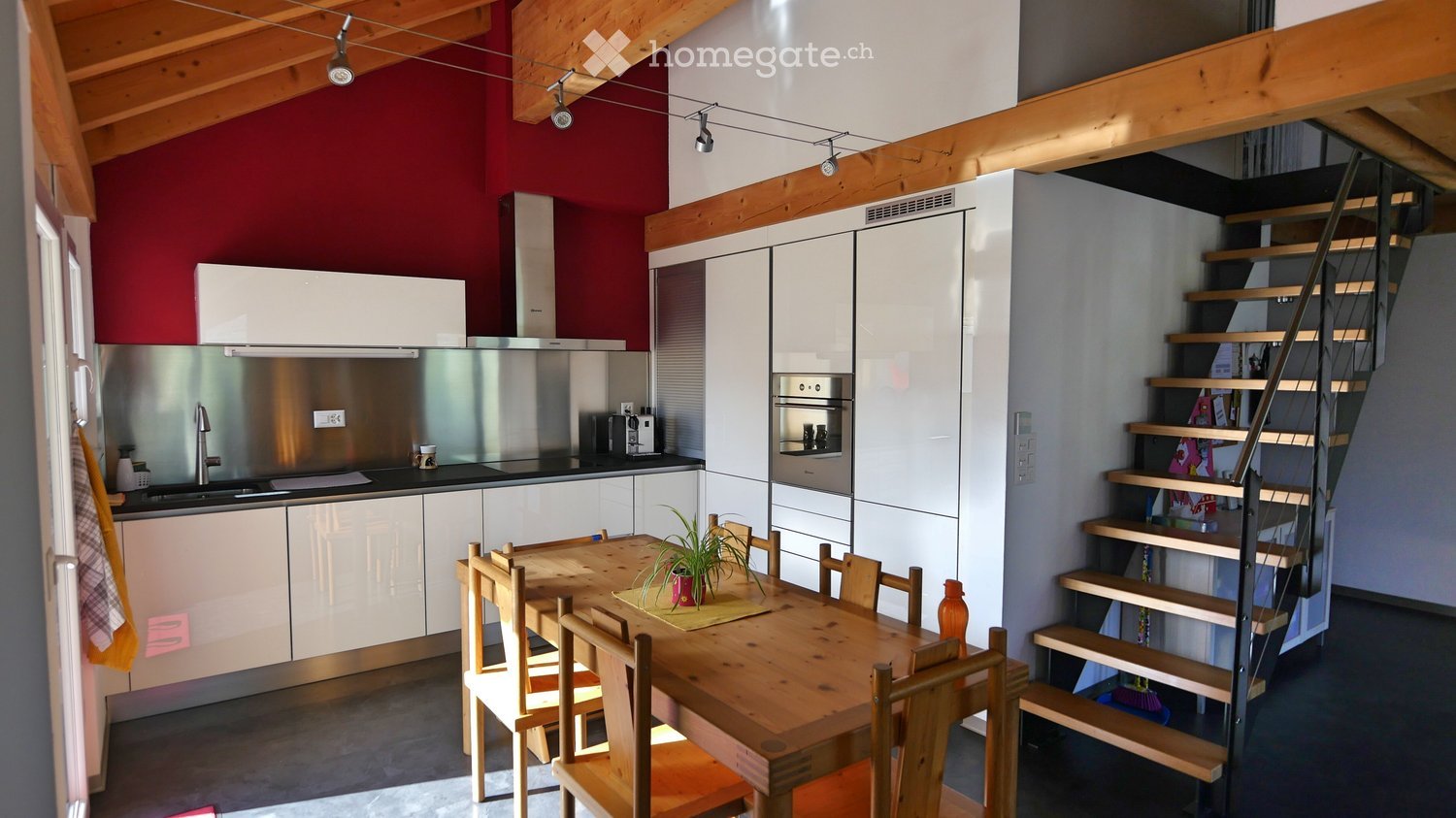 Modern kitchen with white cabinets, stainless steel appliances, a wooden dining table with chairs, and a staircase leading to another level.