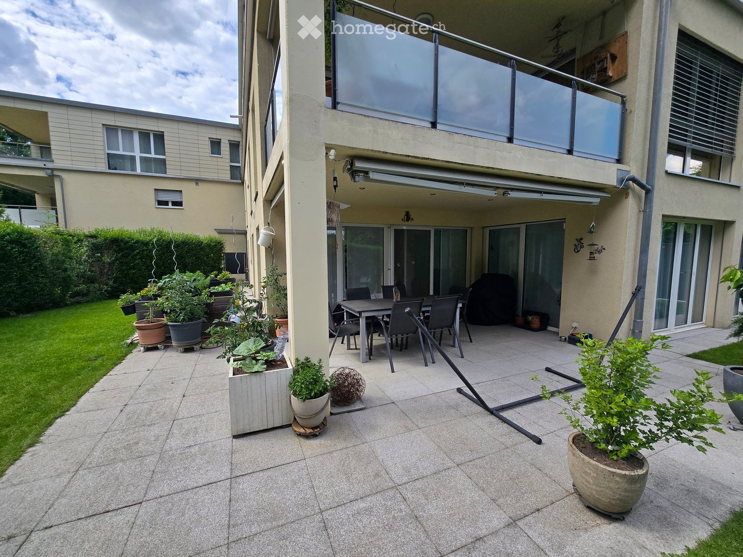 A modern outdoor patio with a tiled floor, a dining table with chairs, potted plants, and a barbecue. The area is connected to a building with a balcony and is surrounded by a well-maintained lawn and hedges.