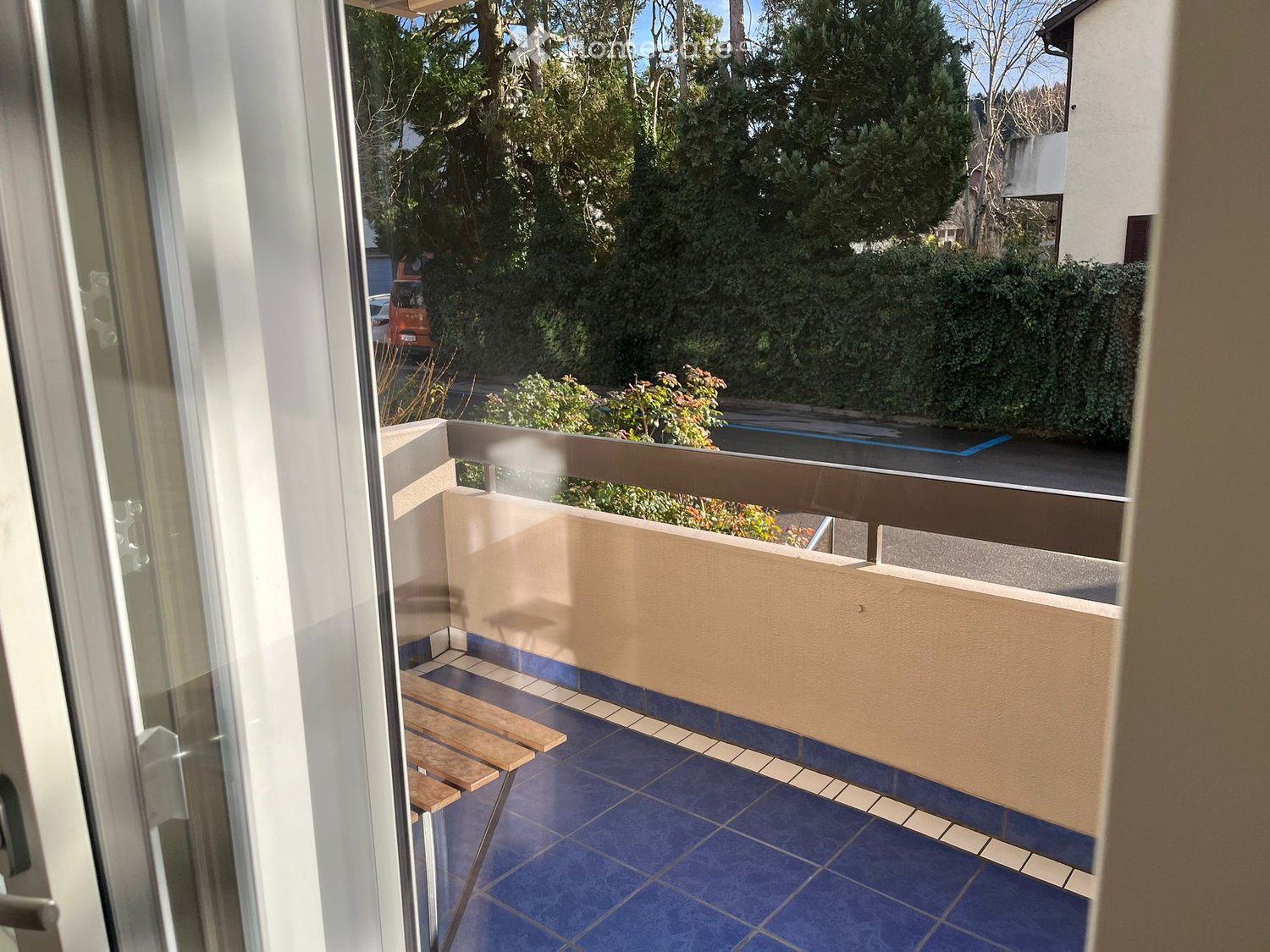 Balcony with blue tiles, wooden bench, and a view of greenery and a parked car.