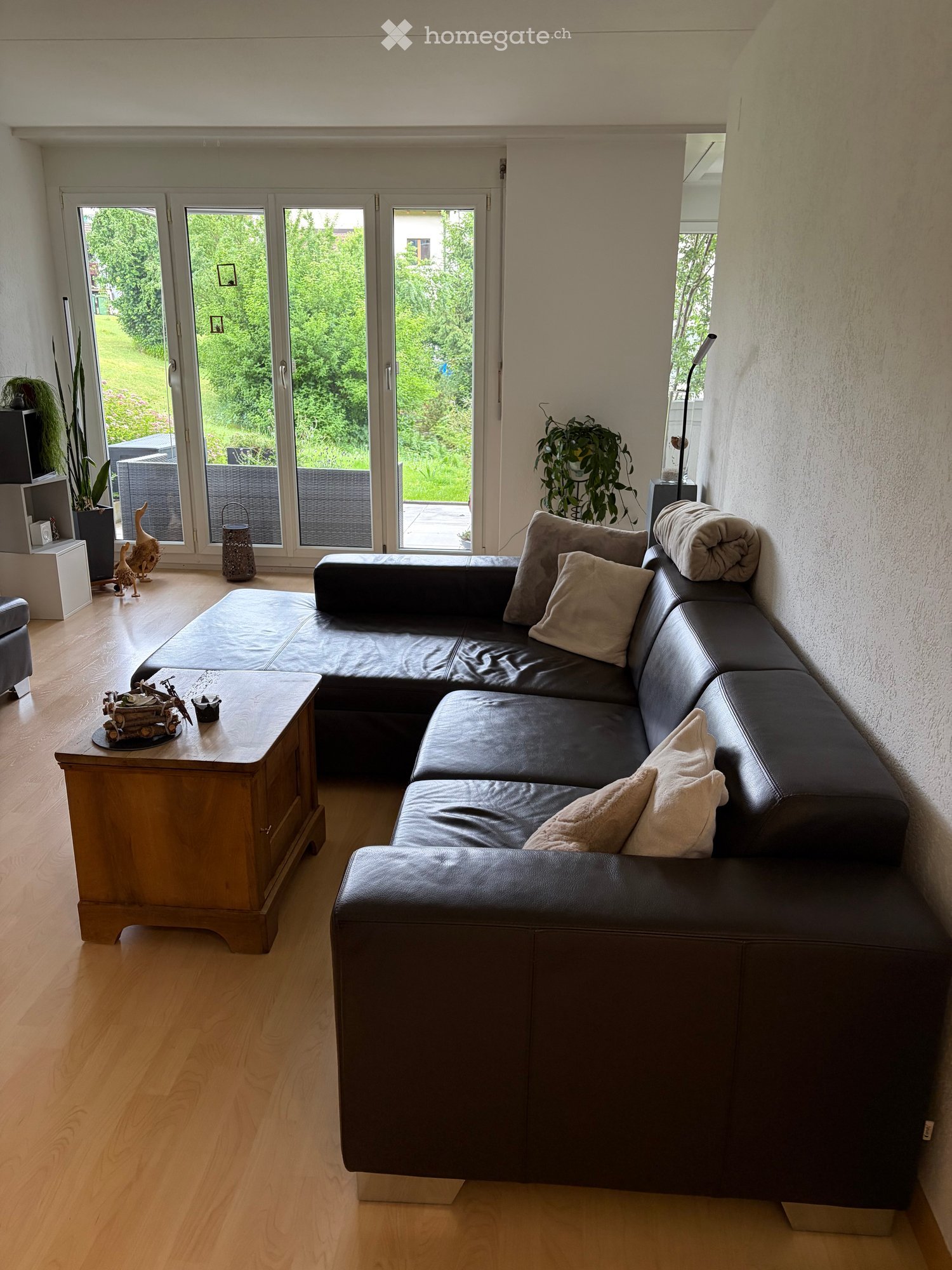 Modern living room with a black leather sectional sofa, wooden coffee table, potted plants, and large windows overlooking a garden.
