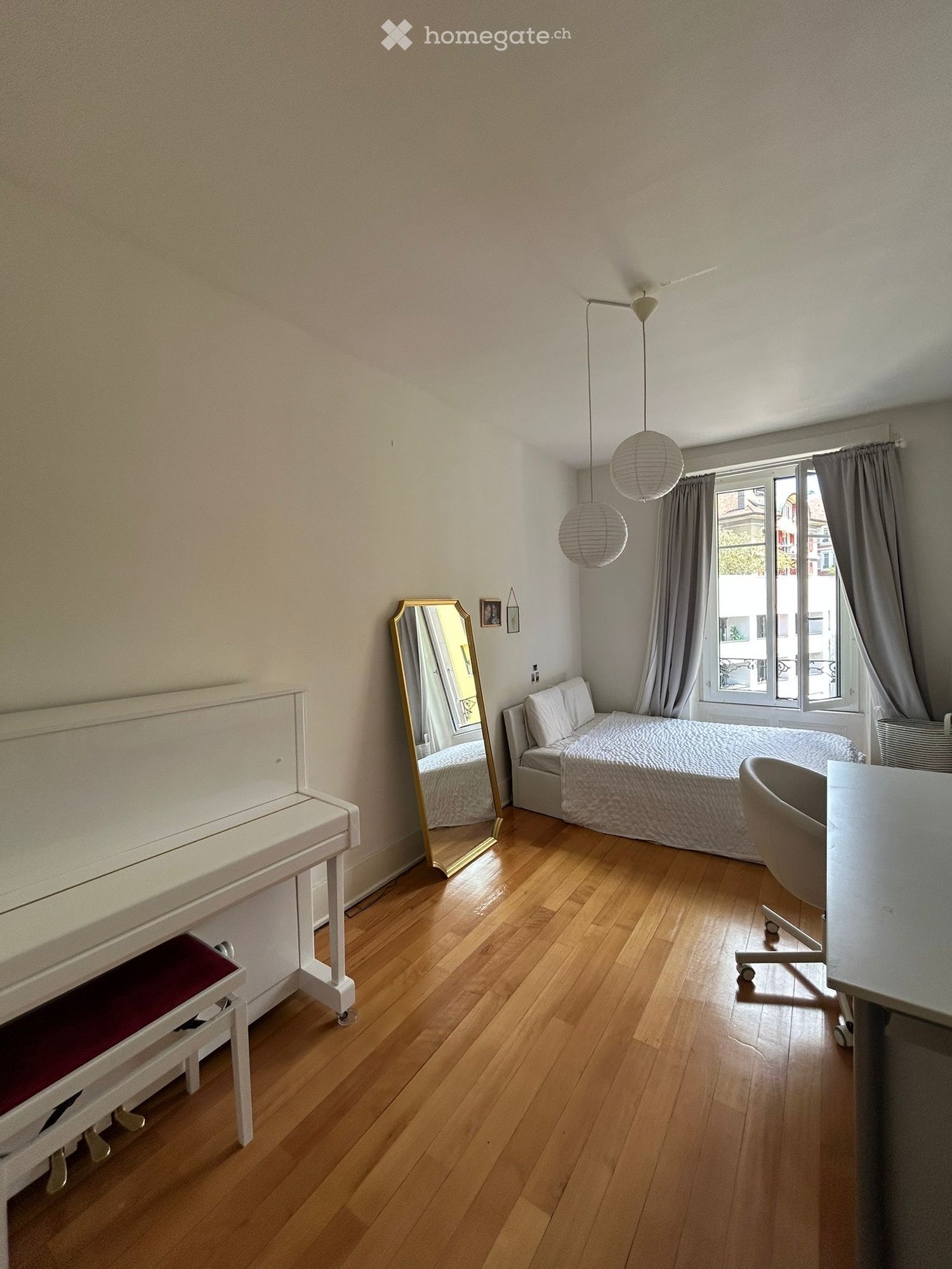 A well-lit bedroom with wooden flooring, a bed with white bedding, a white piano with a red seat, a large mirror, a hanging lamp, and a small table with a chair.