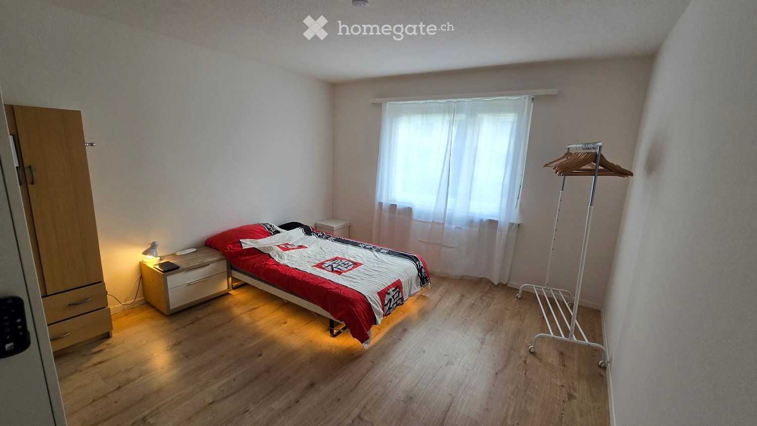 Cozy bedroom with wooden flooring, a single bed with red and black bedding, a nightstand with a lamp, a wardrobe, and a coat rack.