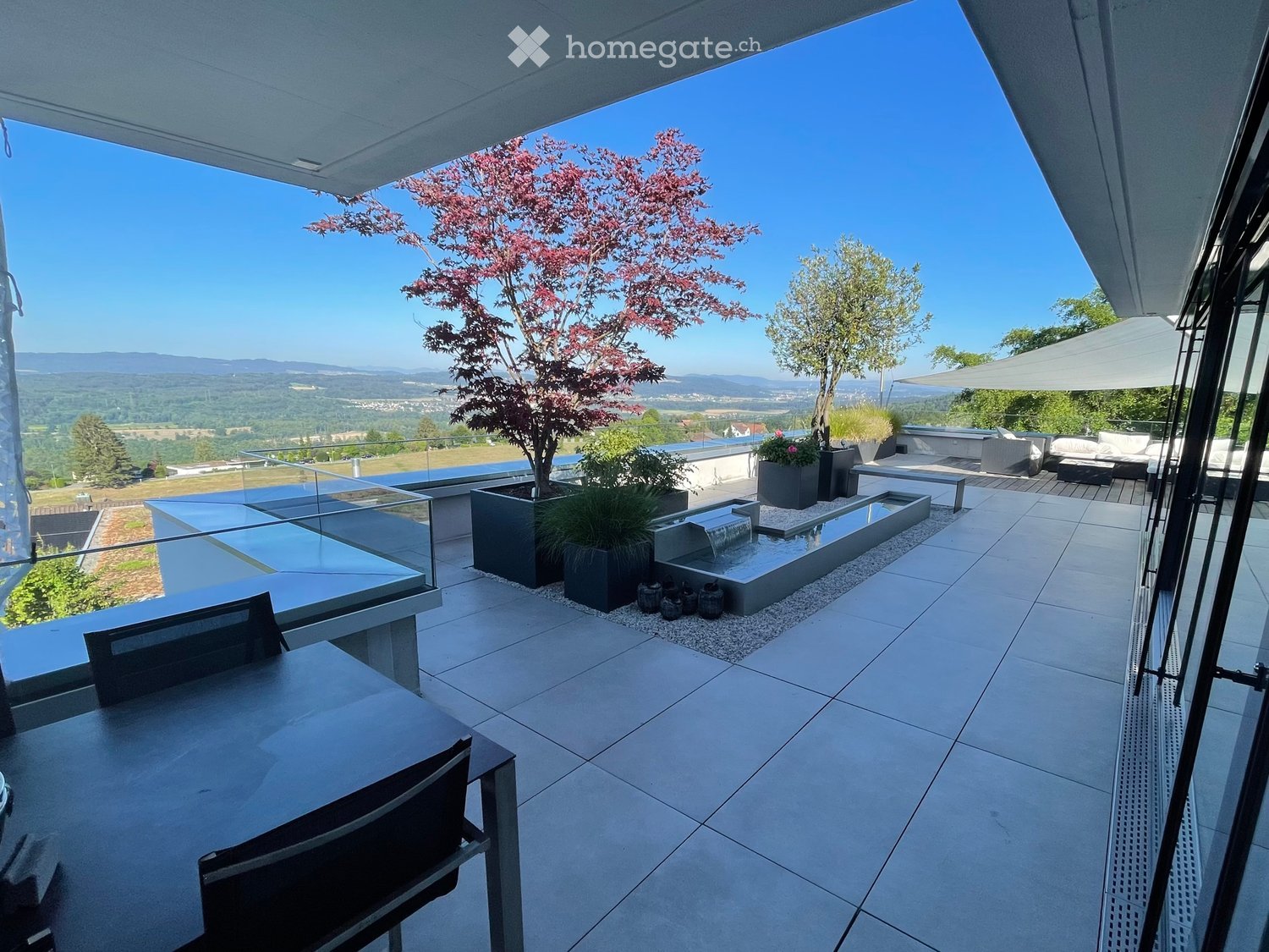 Modern rooftop terrace with tiled flooring, glass railing, water feature, potted trees, and outdoor seating area with a view of the countryside.
