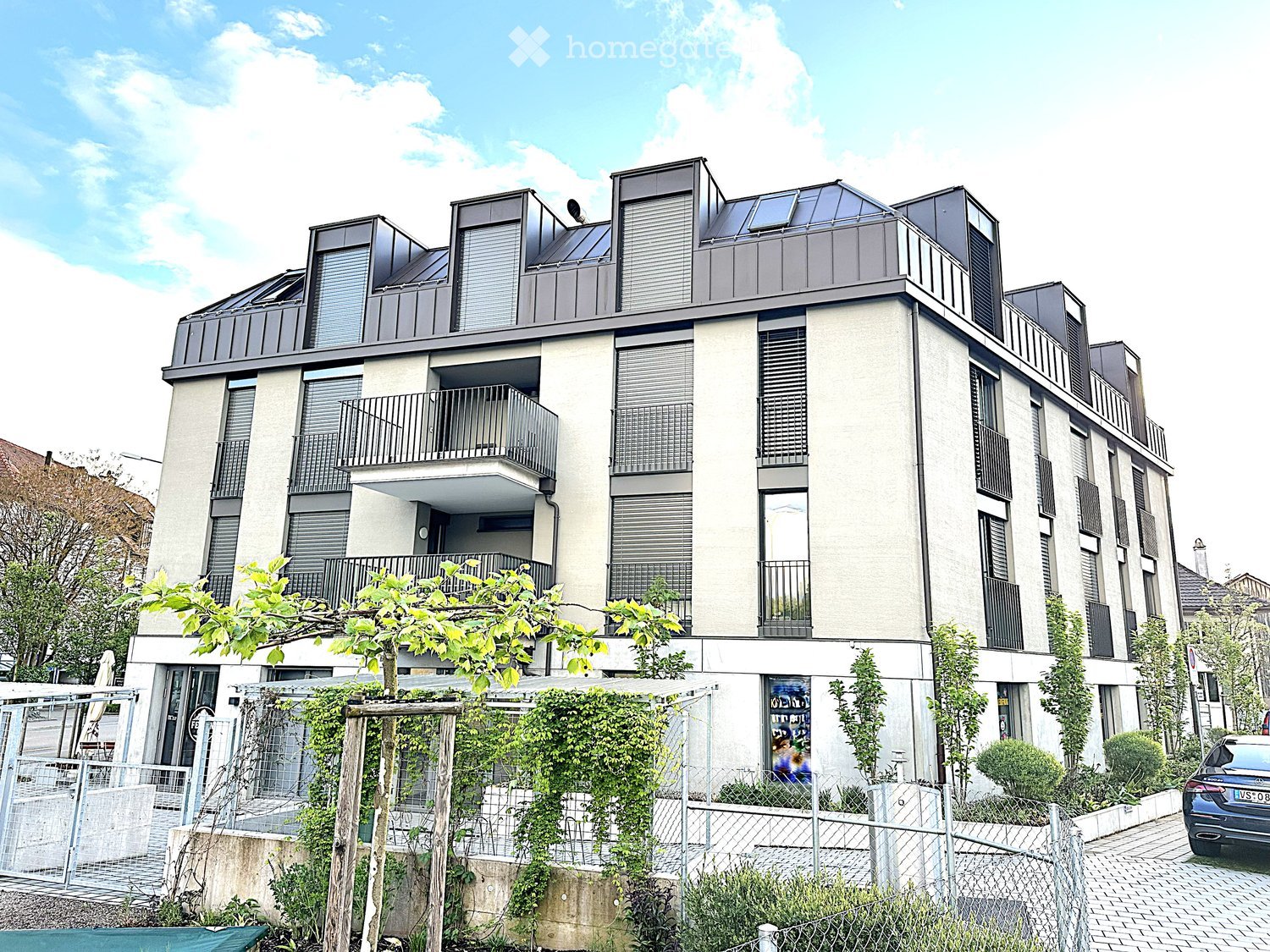 Building. Modern multi-story residential building with balconies, large windows, and a rooftop terrace. The facade is light-colored with dark accents, and the surrounding area includes a small garden and a parked car.