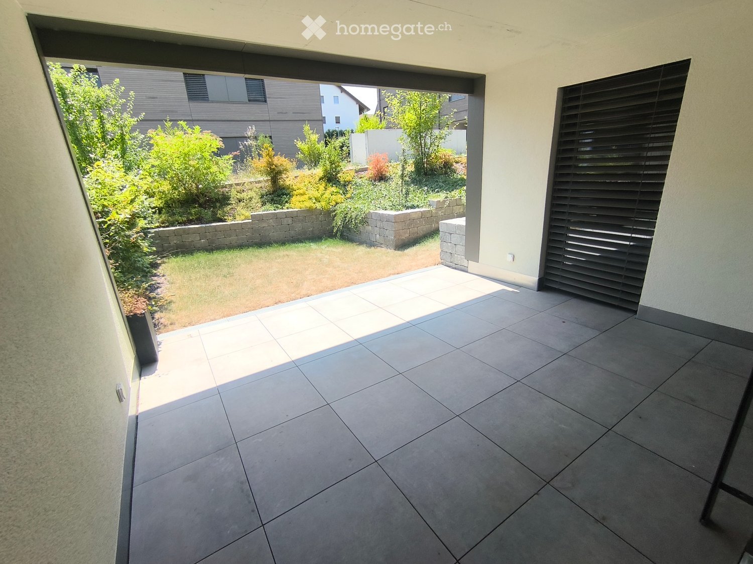 Balcony with tiled floor, large sliding glass door, view of garden with low stone walls and greenery.