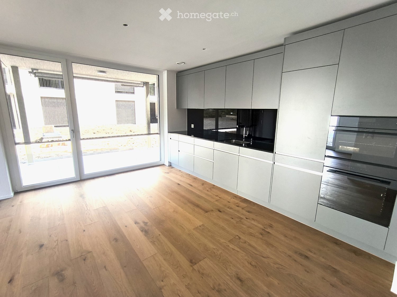 Modern kitchen with light grey cabinets, black countertops, and wooden flooring. Features a large window and sliding glass doors leading to an outdoor area.