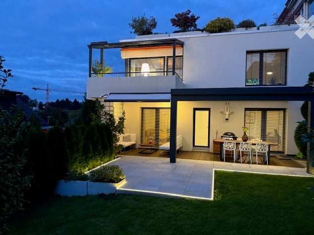 Modern two-story house with a flat roof, large windows, and a spacious outdoor patio area with a dining set and lounge seating.