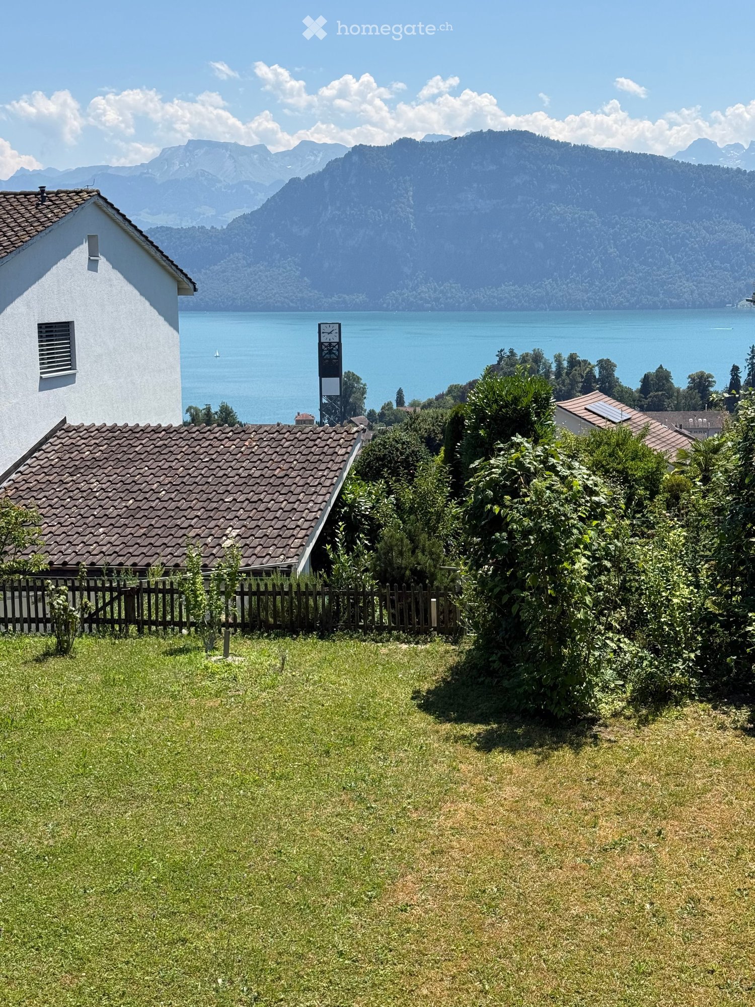Lush garden with a wooden fence, surrounded by greenery, overlooking a serene lake and distant mountains.