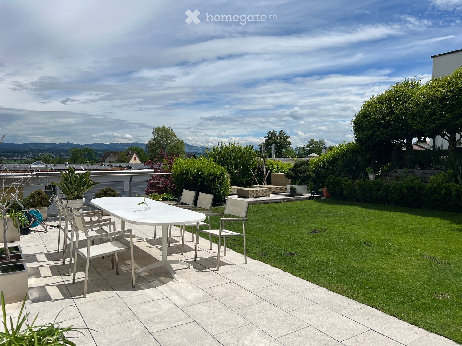 Well-maintained garden with a tiled patio area, white table and chairs, lush green lawn, and various potted plants.