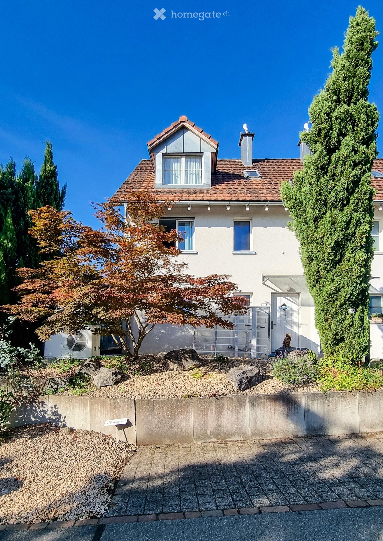 Two-story house with white exterior, red-tiled roof, multiple windows, and a small garden area with trees and rocks.