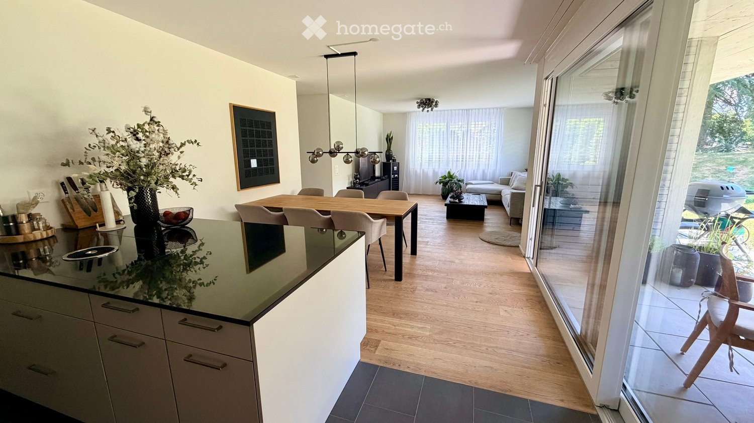 Open-plan kitchen and living area with modern furniture, including a kitchen island, dining table, sofa, and coffee table. Large windows provide natural light and a view of the outdoor space.