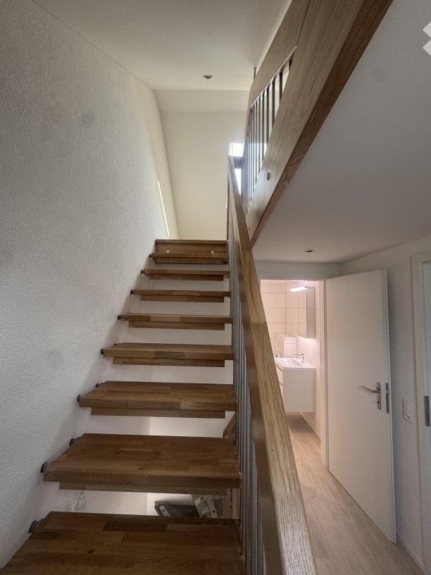 Staircase with wooden steps and a glass railing leading to a bathroom area with white tiles and a sink.