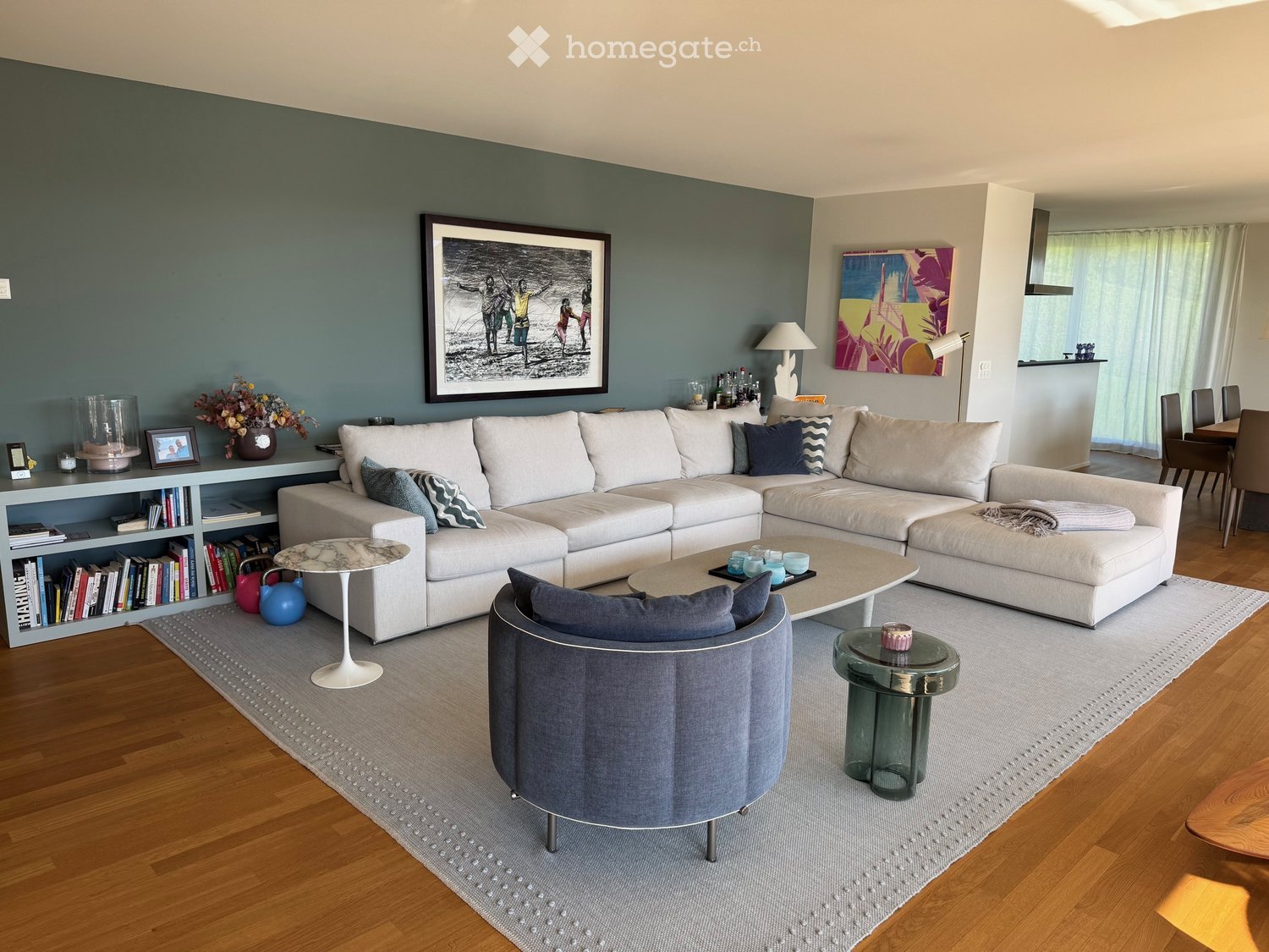 Modern living room with a white sectional sofa, coffee table, armchair, bookshelf, and decorative items. The space features hardwood flooring and large windows with sheer curtains.