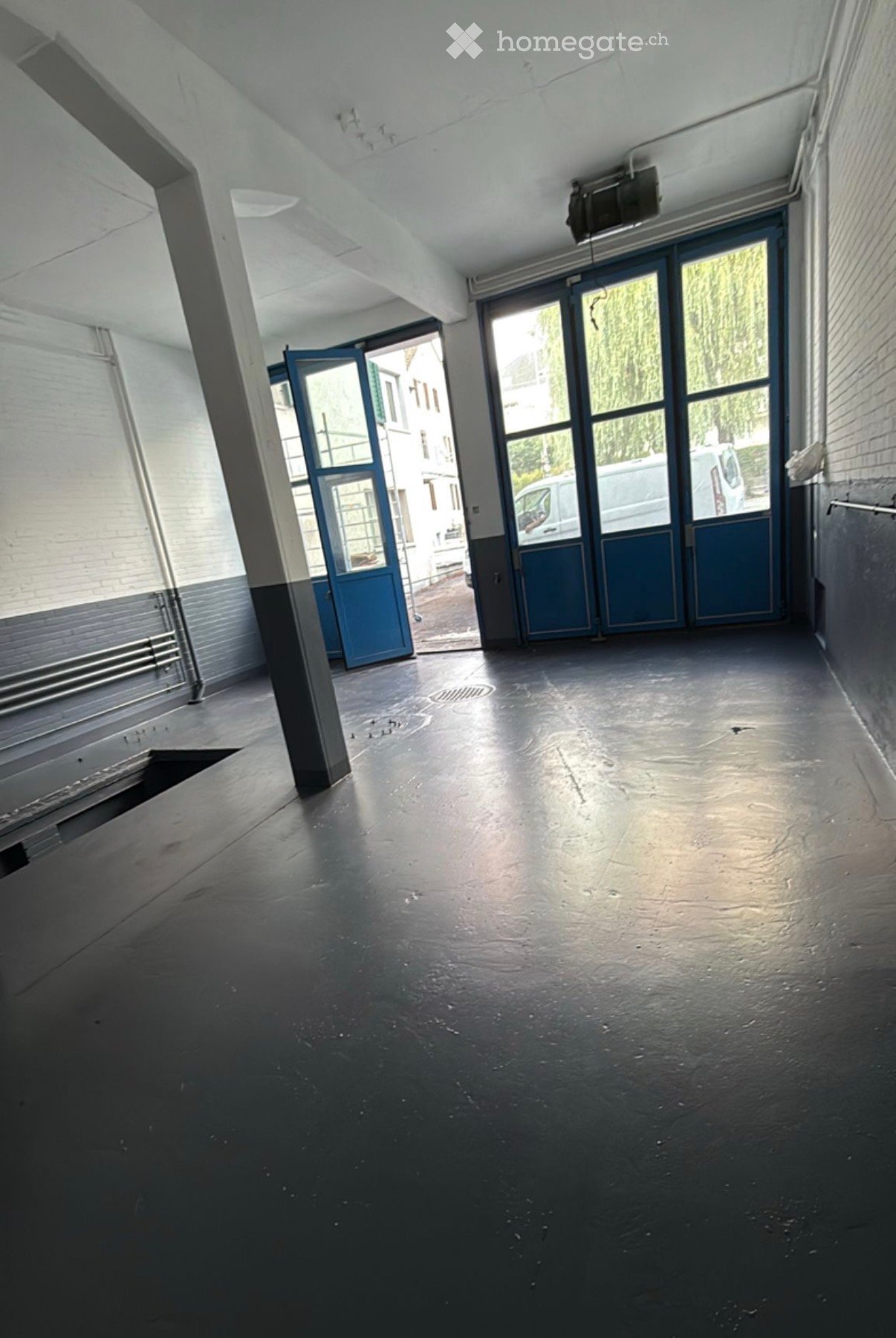 Empty room with dark concrete floor, white brick walls, large windows, and blue doors leading outside.