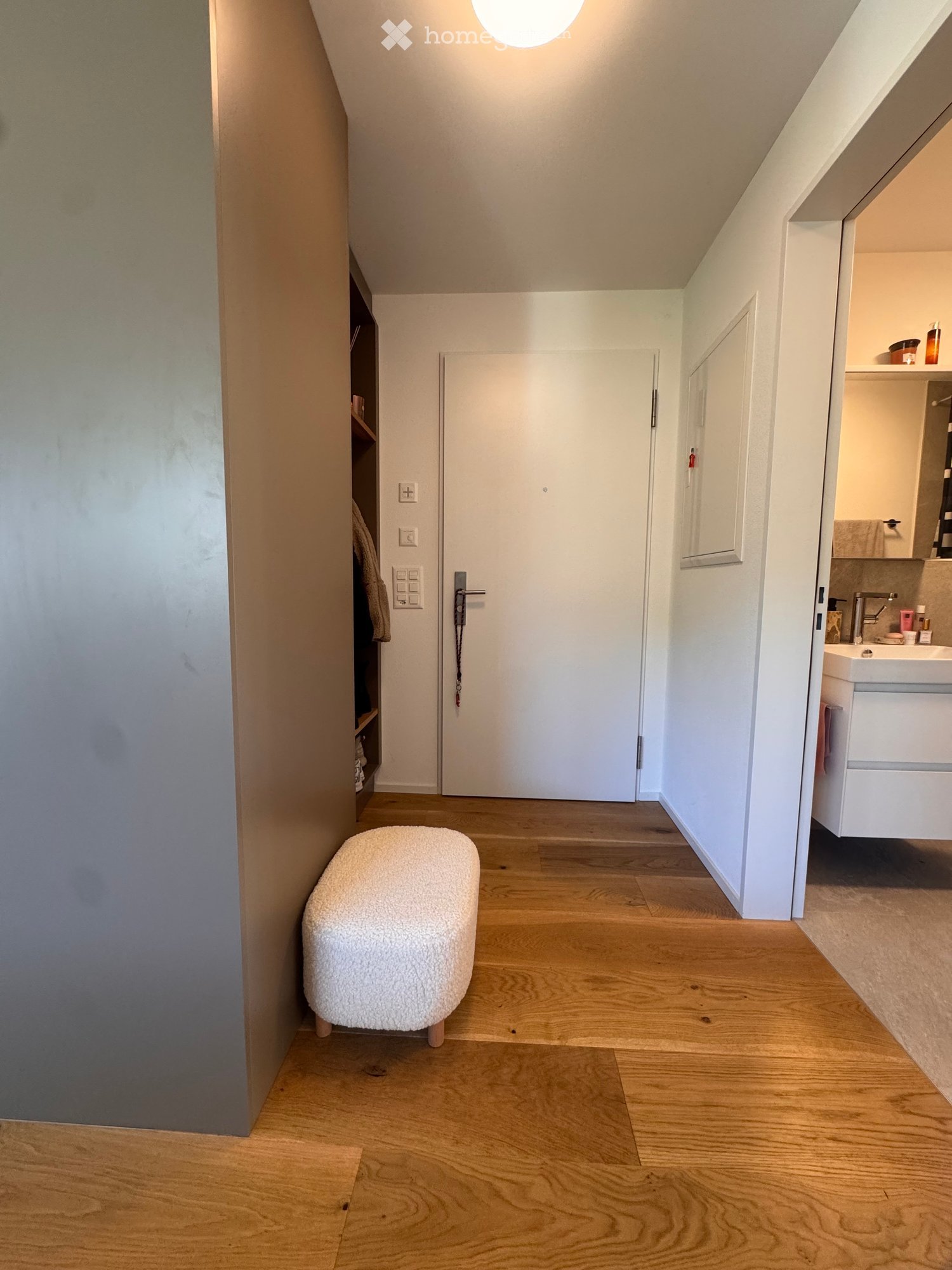 A hallway with wooden flooring, a white door, a small round stool, and a bathroom area with a sink, faucet, and mirror.