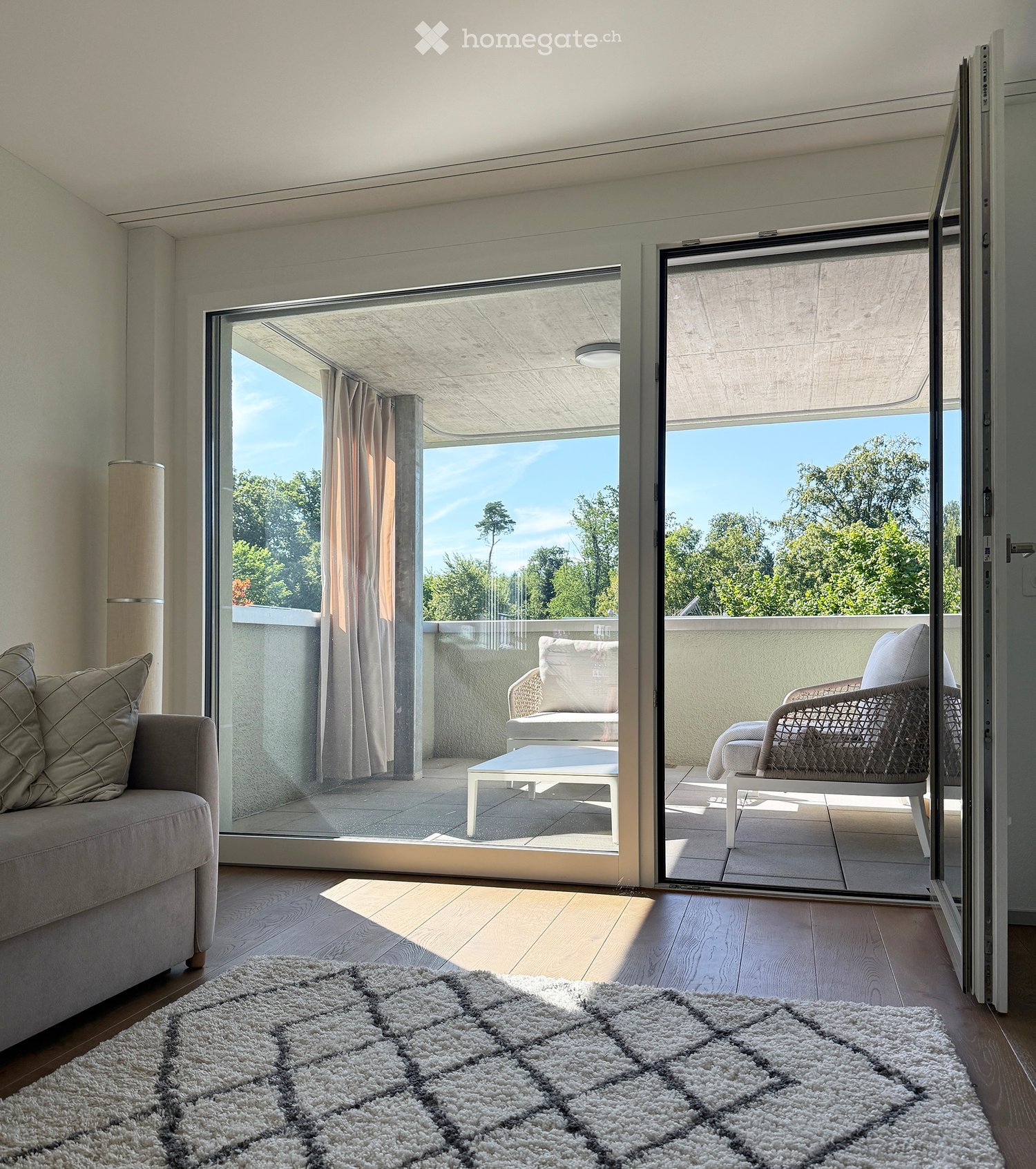 Modern living room with large sliding glass doors opening to a balcony, featuring a grey sofa, white coffee table, and wicker chairs on the balcony.