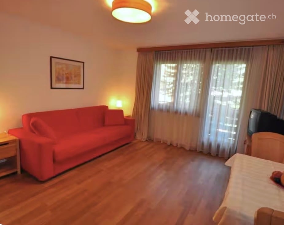 A cozy living room with a red sofa, wooden floor, a small table with a lamp, a television, and a picture frame on the wall. Large windows with curtains let in natural light.